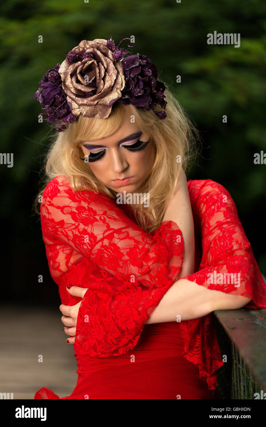 Blonde Modells, feinen Gesichtszügen, purpurrote Blume Krone, geflügelte Eyeliner, riesige Wimpern, rotes Spitzenkleid, kontemplative auf Brücke Stockfoto