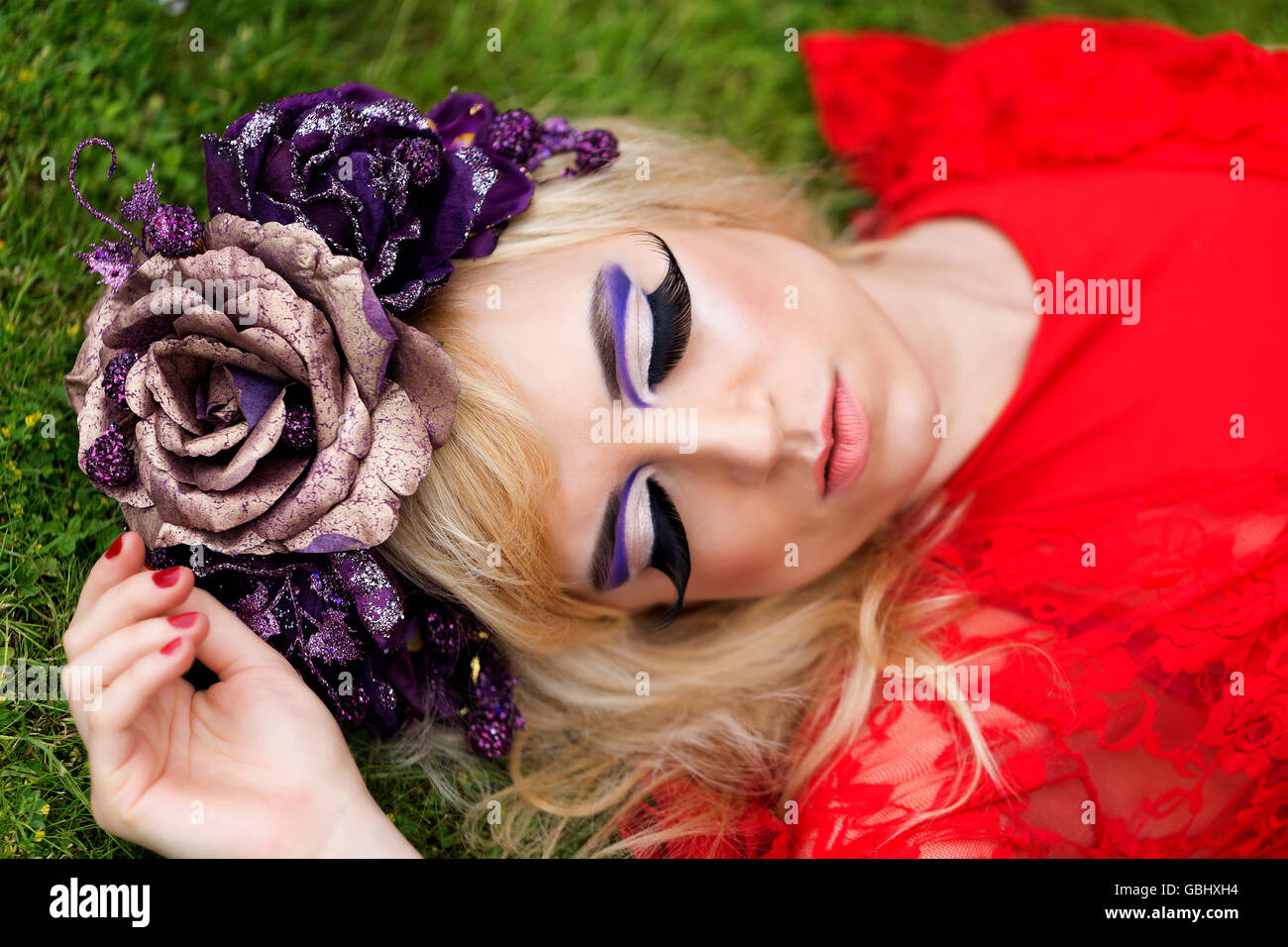 Blonde Modells, feinen Gesichtszügen, purpurrote Blume Krone, geflügelte Eyeliner, riesige Wimpern, rotes Spitzenkleid, schlafen auf dem Rasen liegen Stockfoto