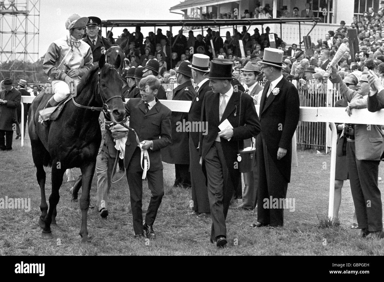 Besitzer-Trainer Arthur Budgett (schwarzer Seidenhut, rechts) schaut stolz darauf, wie sein Pferd Blakeney mit Ernie Johnson nach dem Sieg im Derby Stakes in Führung geht. Stockfoto