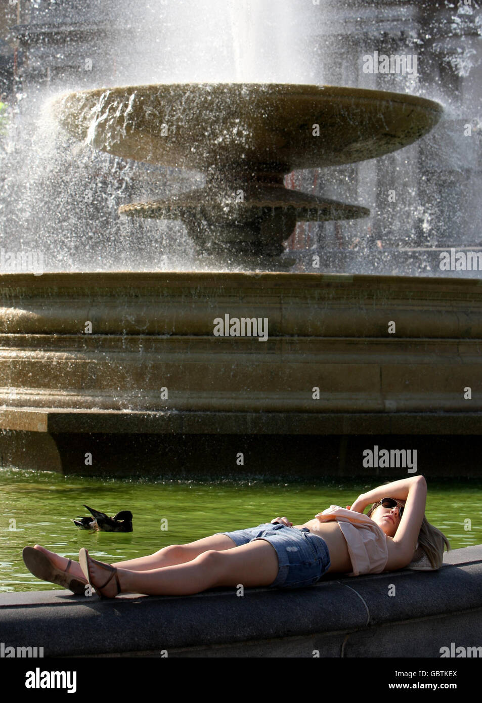 Eine Frau sonnen sich am Rande eines Brunnens am Trafalgar Square im Zentrum von London, der aufgrund einer Algenbildung grünes Wasser hat. Stockfoto