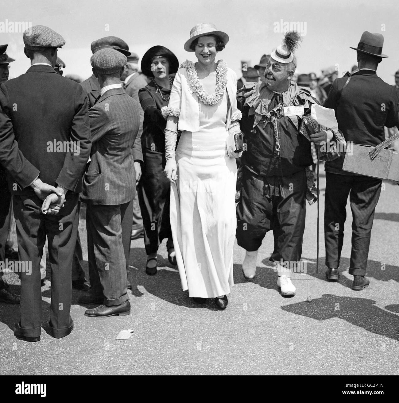 Eine gut gekleidete Dame bei den Rennen in Royal Ascot zeigt ihre Belustigung, als sie von einem Clown in vollem Kostüm begleitet wird, während sie sich durch die Menge macht. Stockfoto