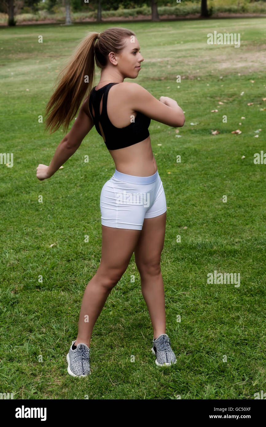 Attraktive kaukasischen Teen Girl Doing verdrehen Übungen In weißen Hosen und schwarzen Top stehen auf dem grünen Rasen im Park Stockfoto