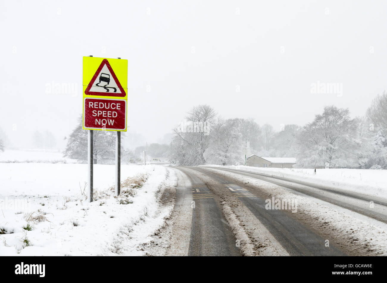 Eine rutschige Oberfläche Warnung Verkehrszeichen auf der Seite der Landstraße während es schneit mit Schnee auf dem Boden. Stockfoto