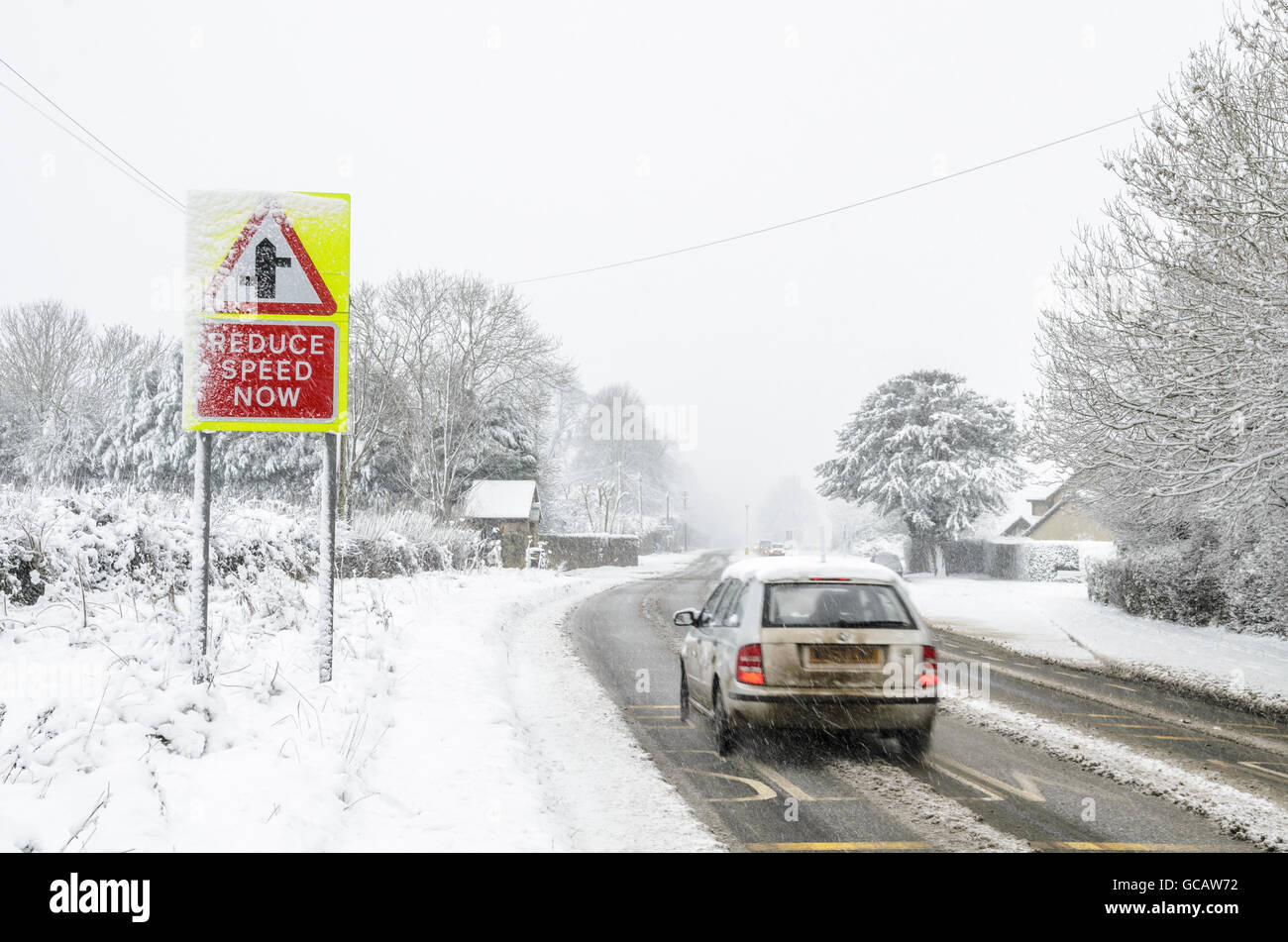 Autos fahren vorbei an einer Straße Zeichen Warnung vor einer Kreuzung voraus während es mit Schnee auf dem Boden schneit. Stockfoto