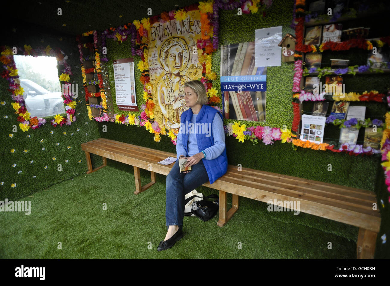 Eine Frau liest, während sie auf einen Bus in einer Bushaltestelle wartet, die beim Hay Festival in Hay-on-Wye mit Plastikblumenkränzen und Astro-Rasen in eine Buchhandlung umgewandelt wurde. Stockfoto