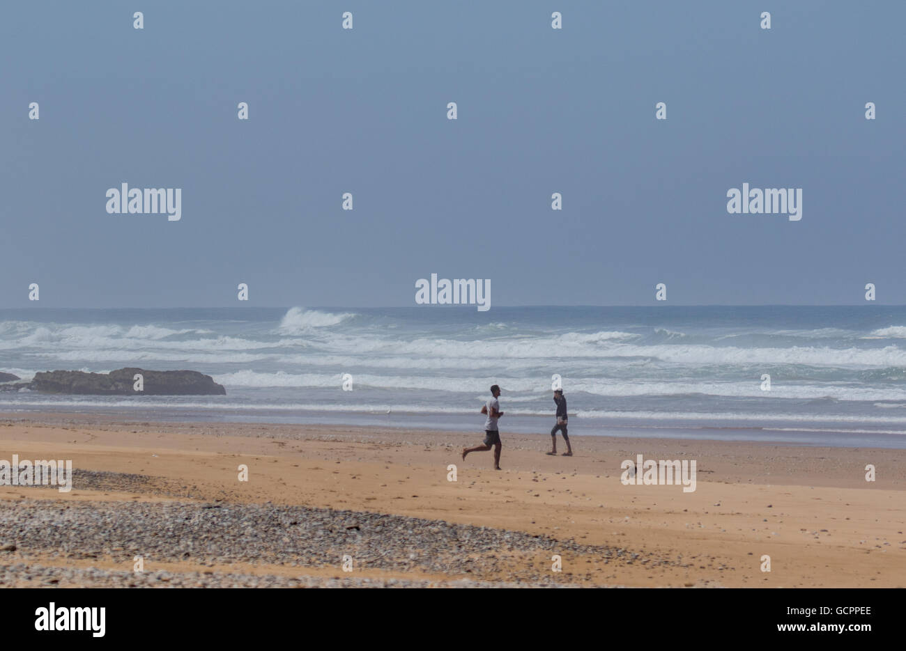 Zwei junge Männer spielen Fußball auf einem Strand mit Wellen im Hintergrund. Stockfoto