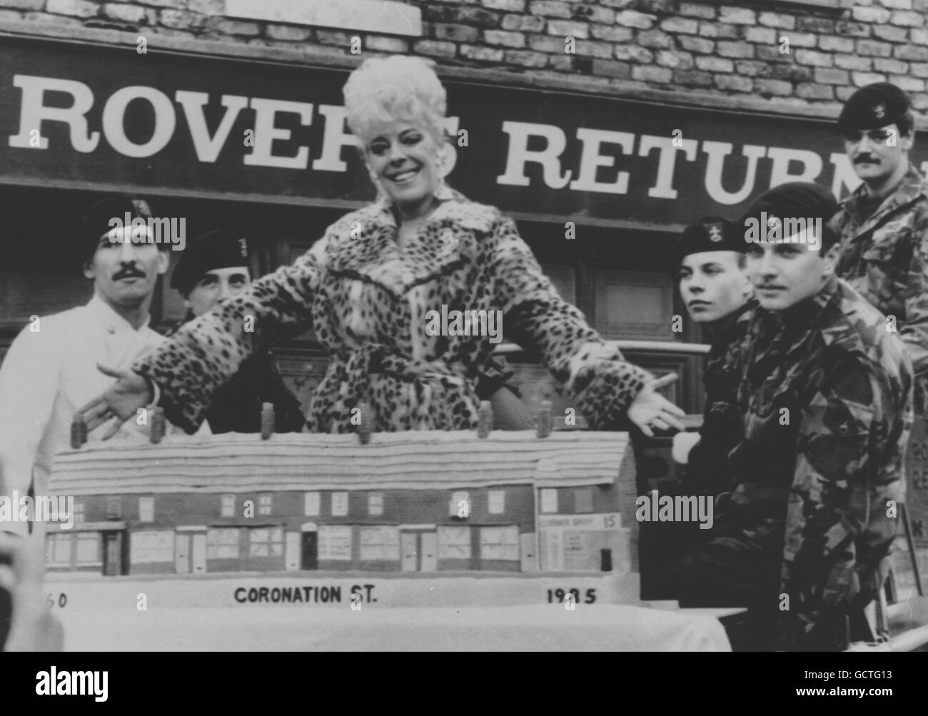 Schauspielerin Julie Goodyear nimmt eine Nachbildung des 'Street' in Form eines Kuchens entgegen, der vom Armeekoch CPL Ron Davies, links, vom Kings Regiment, TA Unit, gebacken wurde. Der Kuchen wird im Dorchester Hotel in London gegessen, wenn die Darsteller den Silbernen Jahrestag der Seifenoper feiern. Stockfoto