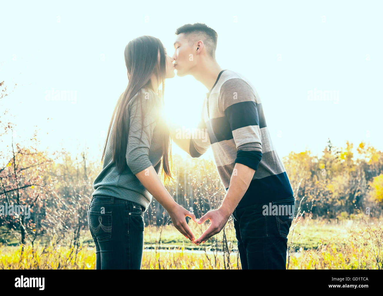 Eine junge asiatische paar küssen in einem Park im Herbst und ein Herz mit ihren Händen in die Wärme der untergehenden Sonne; Edmonton, Alberta, Kanada Stockfoto