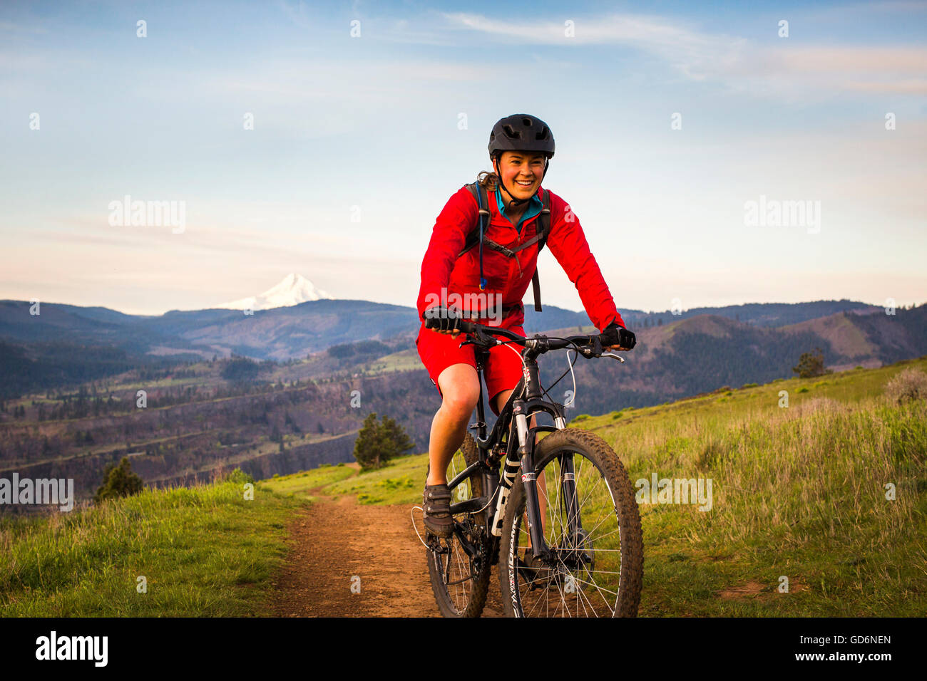 Eine junge Frau in einem roten Mantel reitet ein Mountainbikes auf eingleisige Strecke durch grüne Rasen im frühen Morgenlicht. Stockfoto