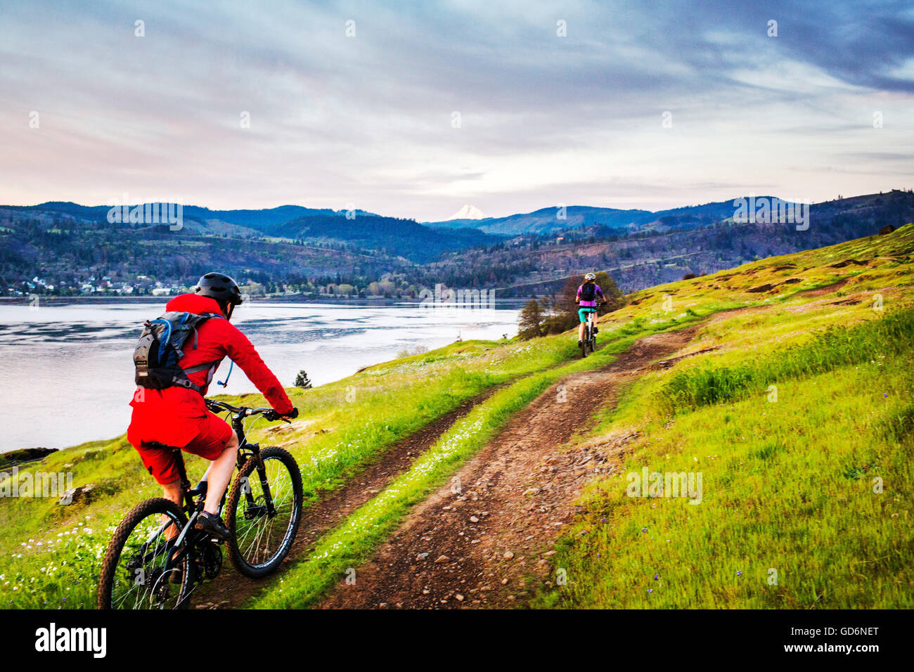 Eine junge Frau im roten Mantel reitet ein Mountainbike auf eingleisige Strecke durch grünen Rasen mit einem großen Fluss und Bergen im Abstand. Stockfoto