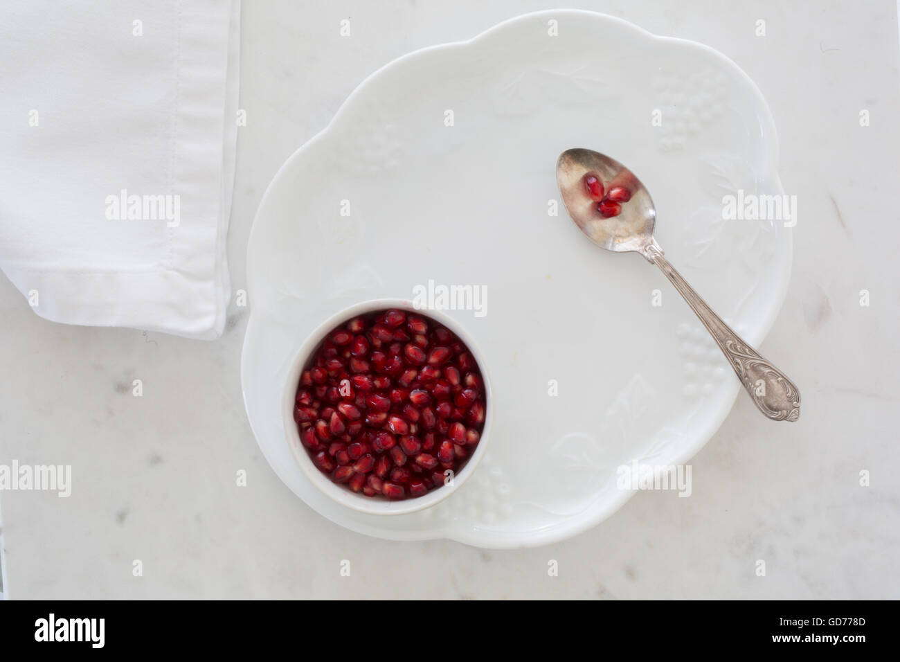 Top-Down Ansicht der Granatapfel Samen in einer Schüssel gesetzt auf einem Dessertteller auf weißem Marmor mit Serviette und Silberlöffel abgespeckt. Stockfoto