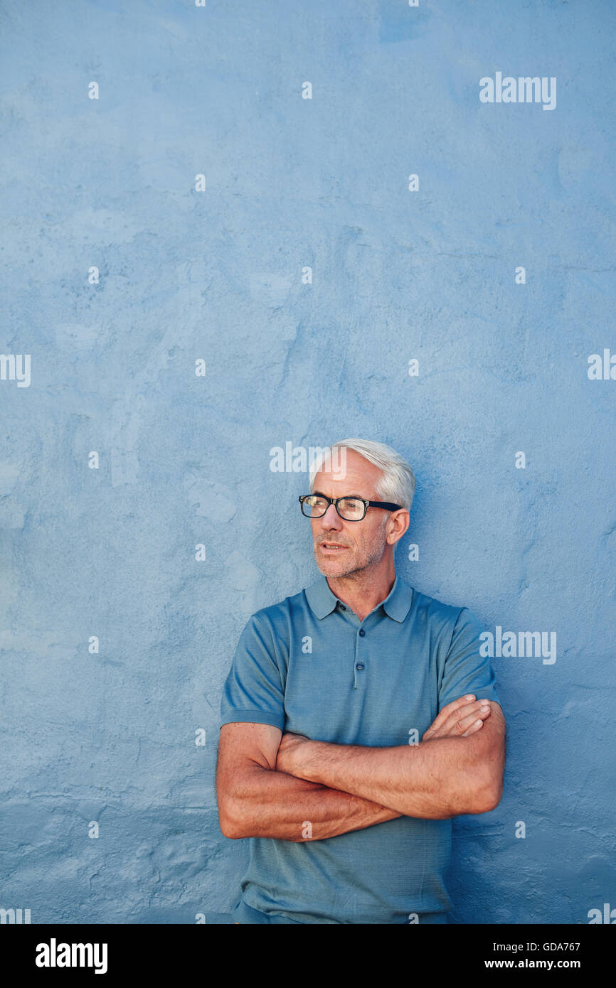 Vertikale Porträt von schöner reifer Mann stehend mit seinem verschränkten Armen und wegschauen vor blauem Hintergrund. Stockfoto