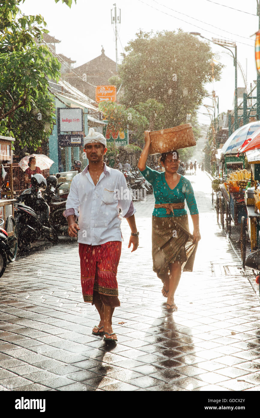 Indonesische Mann und Frau in traditioneller Kleidung zu Fuß unter dem Regen auf der Straße von Kuta, Indonesien Stockfoto