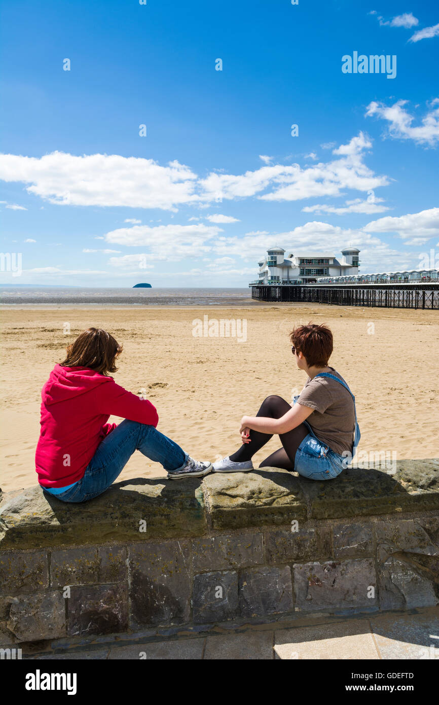 Zwei junge Frauen sassen auf der Promenade an der Wand mit Blick auf den Strand und das Grand Pier in Weston-super-Mare, Somerset, England. Stockfoto