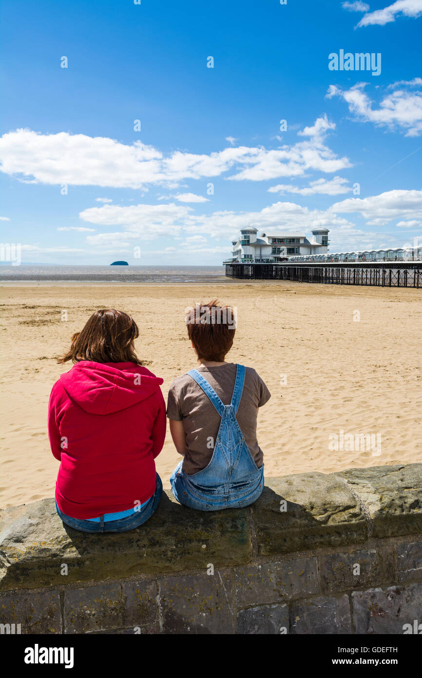 Zwei junge Frauen sassen auf der Promenade an der Wand mit Blick auf den Strand und das Grand Pier in Weston-super-Mare, Somerset, England. Stockfoto