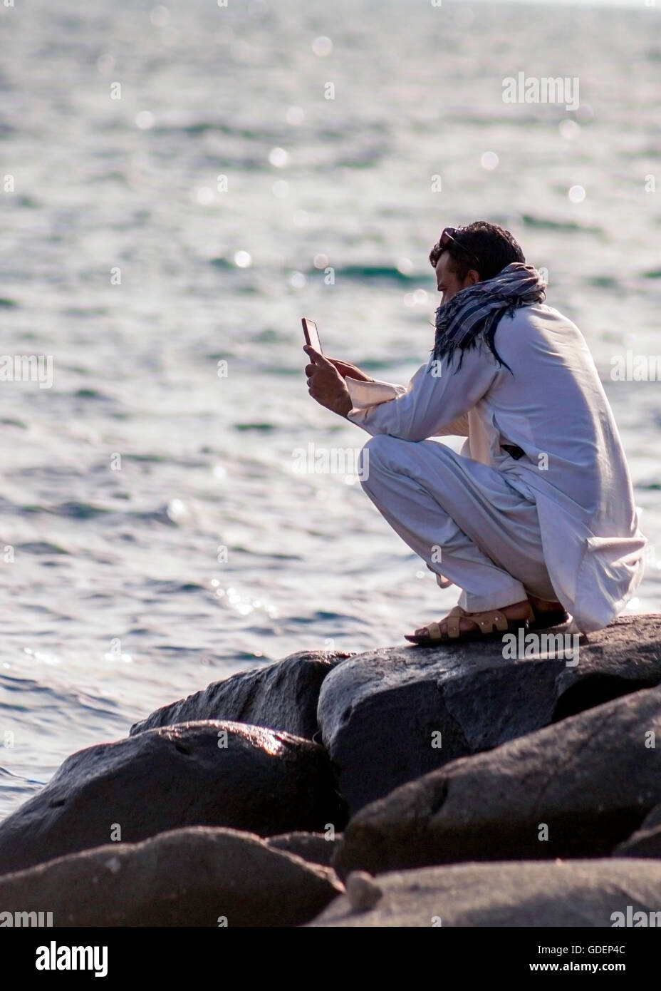 Mann sitzt auf Felsen mit einem Handy am Meer / Strand - Rotes Meer Stockfoto