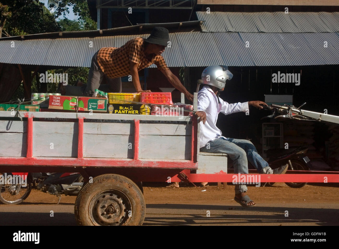 Ein Mann fährt ein Motorrad, das Ziehen eines Anhängers, beladen mit Lebensmitteln & Getränk Elemente auf einer Stadtstraße in Chork Village, Kambodscha. Stockfoto
