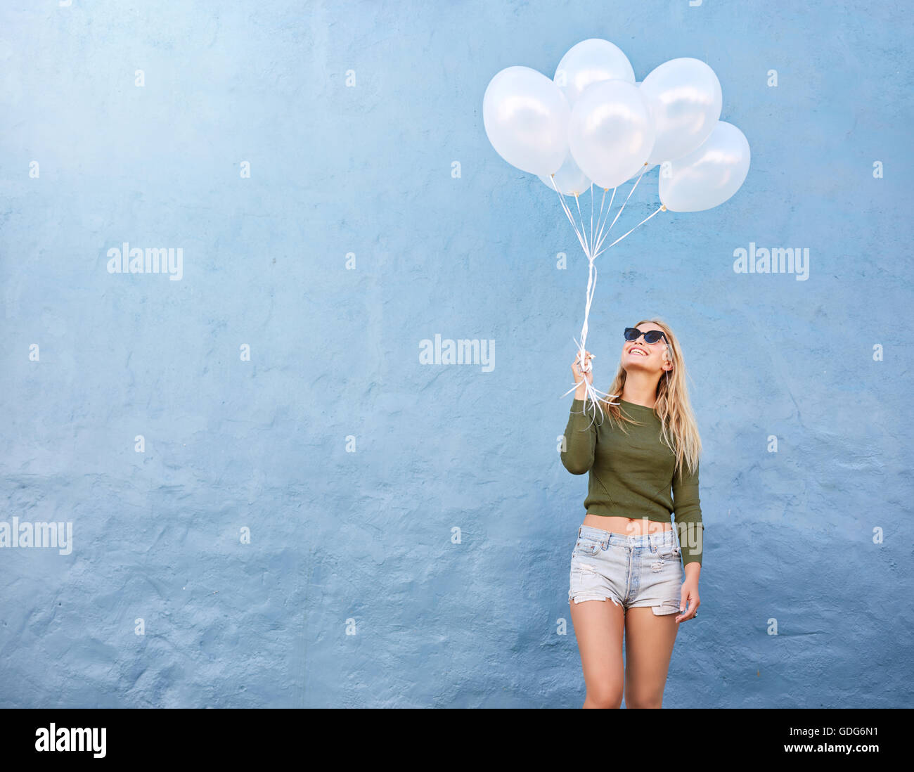 Schuss der glückliche junge Frau mit Haufen Luftballons gegen blauen Wand. Attraktives weibliches Modell in stilvollen Casuals halten weiße bal Stockfoto