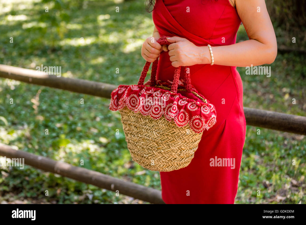 weibliche Hände halten eine Tasche im Landhausstil hergestellt aus Bast und rotes Tuch mit weißen Blüten, wartet die Frau, gekleidet in einen roten Etuikleid nervös in einem park Stockfoto
