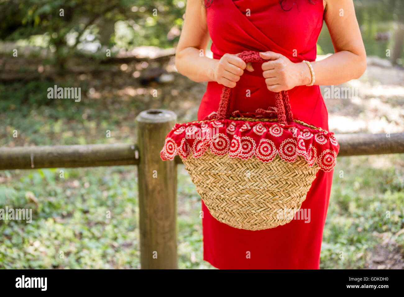 weibliche Hände halten eine Tasche im Landhausstil hergestellt aus Bast und rotes Tuch mit weißen Blüten, wartet die Frau, gekleidet in einen roten Etuikleid nervös in einem park Stockfoto