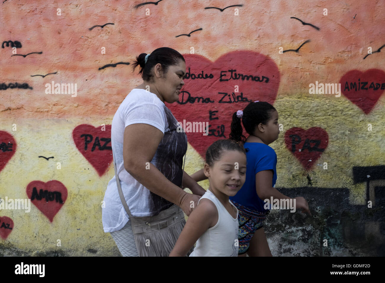 Eine Familie Spaziergänge vorbei an eine Wand gemalt von Friedensaktivisten Marieluce in der Favela Complexo Alemão in Rio De Janeiro, Brasilien, 16. Juli 2016. Die Complexo ist einer großen Favela in Rio De Janeiro, bestehend aus 25 Siedlungen. Marieluce setzt sich die Häuser mit ihren bunten Bildern verschönern. Sie hat bereits rund 40 Hausfassaden mit roten, blauen und gelben Häuser sowie fliegende Drachen als Symbol für das vielfältige Leben in der Favela gemalt. Sie möchte auch halten den Jugendlichen, ihre Farbe helfen der Häuser, um sie zu schützen, das schlechte und Verkauf von Drogen wie die vielen Banden beschäftigt Stockfoto