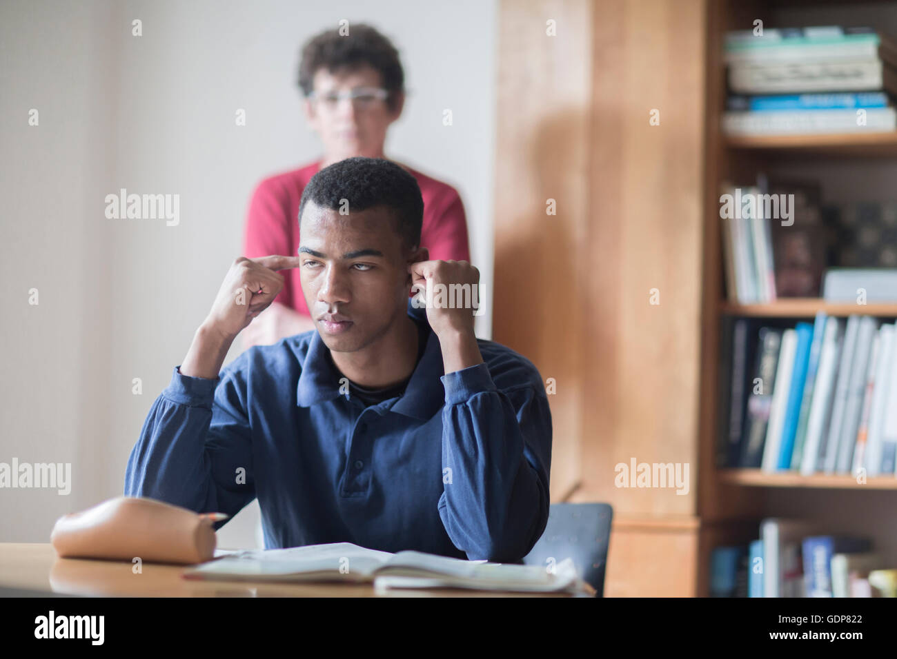 Junge männliche High-School-Schüler sitzen am Schreibtisch mit den Fingern in den Ohren und Lehrer stehen hinter Stockfoto