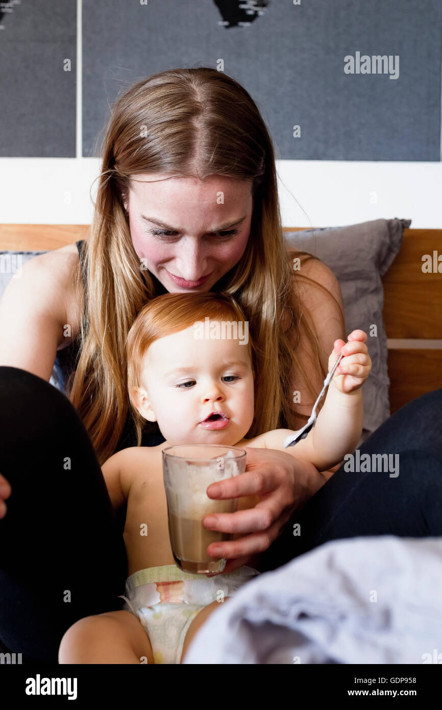 Babymädchen und Mutter im Bett spielen mit Teelöffel und Glas Kaffee Stockfoto