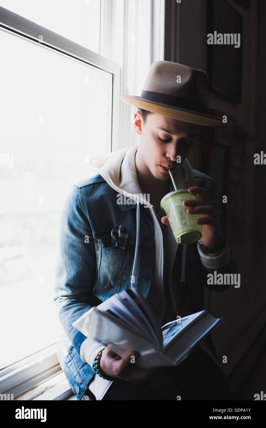 Junger Mann zu Hause, sitzt auf der Fensterbank, Trinken Gesundheit trinken, Buch lesen Stockfoto