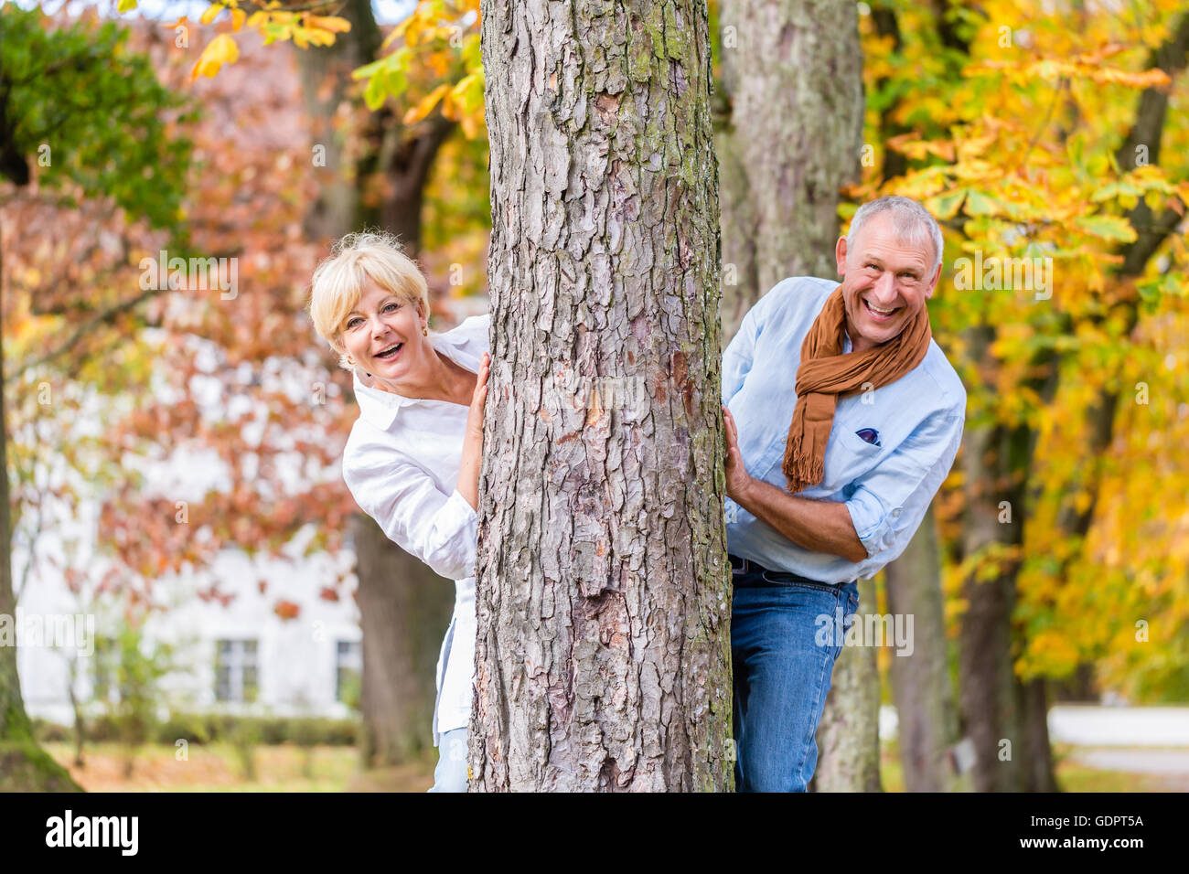 Paar, senior Mann und Frau, flirten mit einander spielen verstecken und suchen, um einen Baum im Herbst Baum Stockfoto