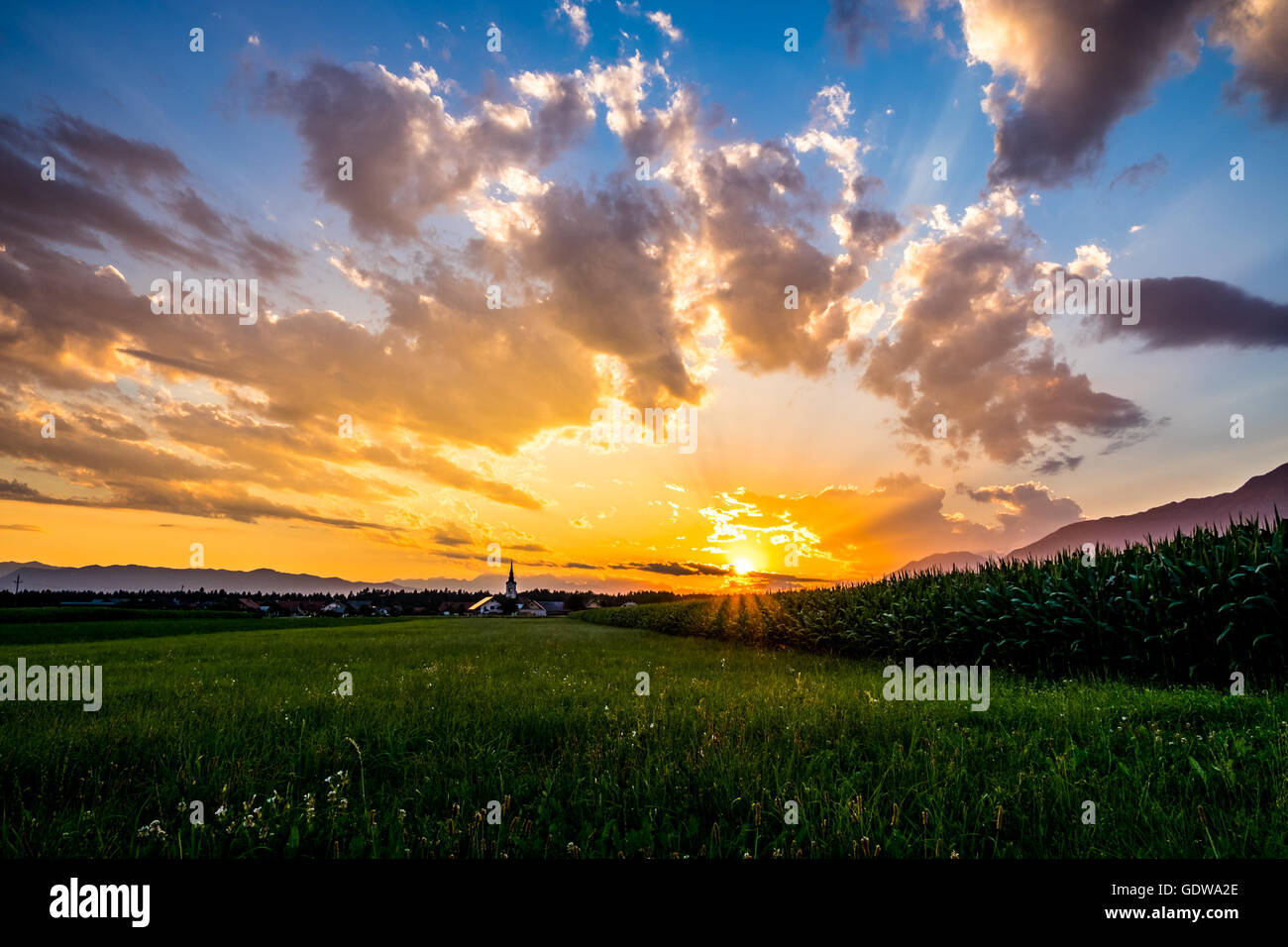 Sonnenuntergang auf dem Lande - dramatischer Himmel mit Wolken, grüne Wiese und Maisfeld, Häuser mit Kirche und Berge auf Zeitmessung Stockfoto