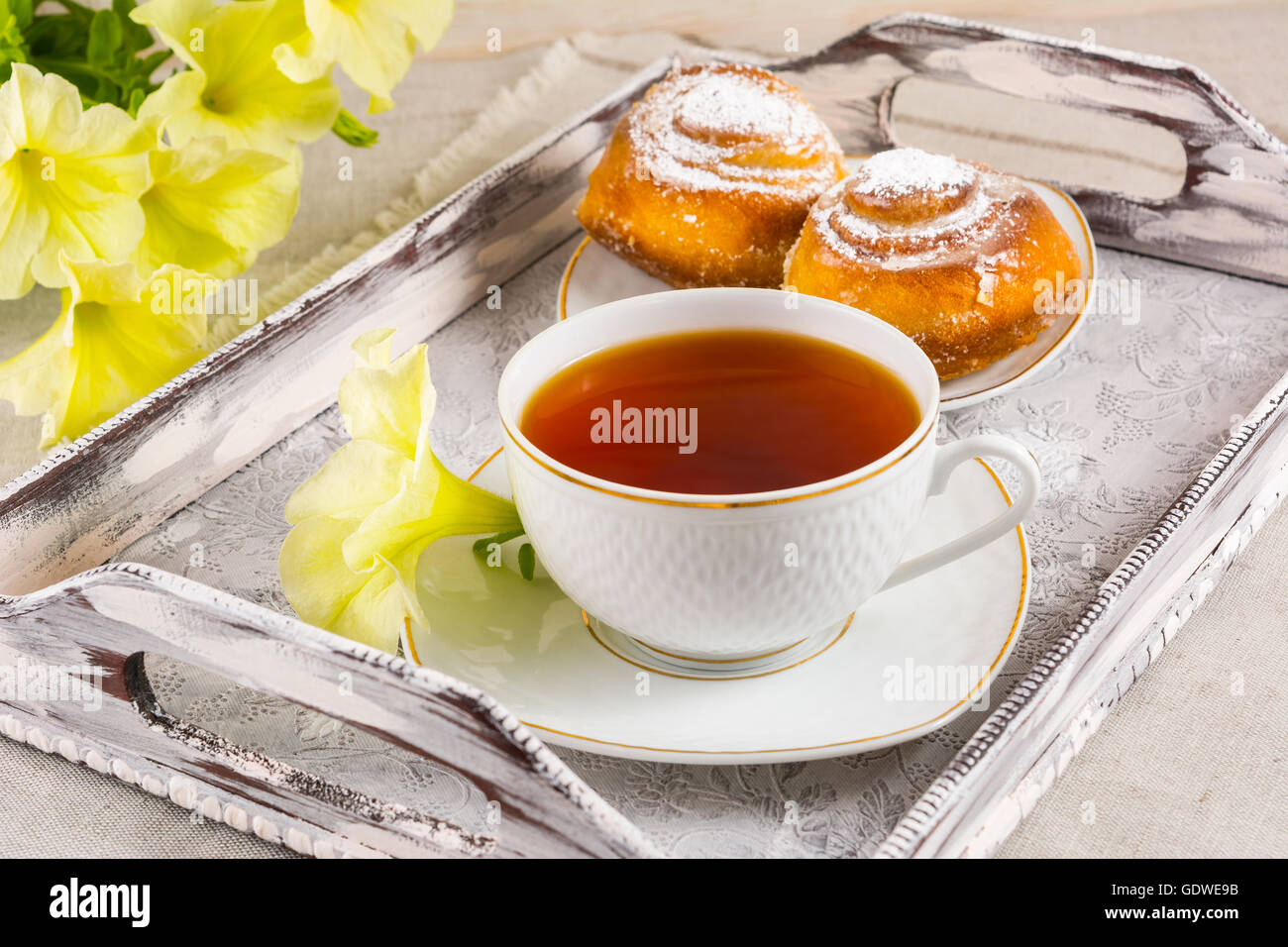 Süße Zimt Brötchen Brötchen und Tasse Tee auf Vintage Serviertablett. Frühstück Plundergebäck und Teetasse mit gelben Blüten. Stockfoto