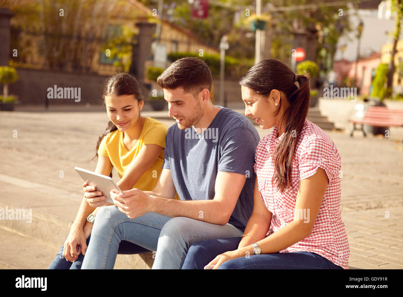 Drei glückliche junge Menschen sitzen in einer Stadt Platz auf einer Stufe mit einem Tablet suchen, während lächelnd lässig in Jeans und t gekleidet-sh Stockfoto
