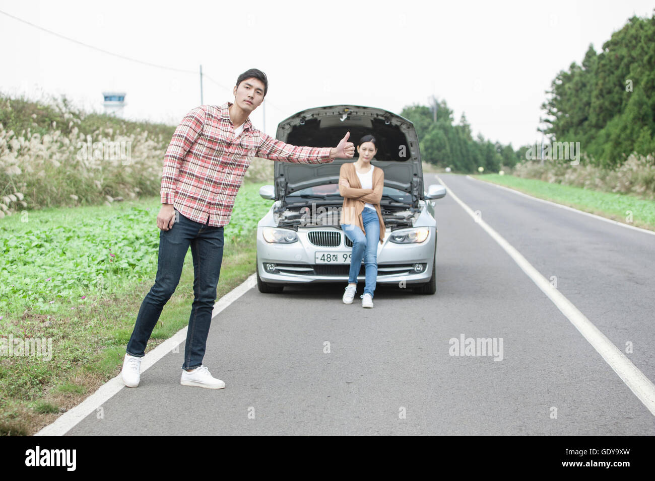 Junger Mann stehen und versuchen, mit einer Frau auf ihr kaputtes Auto sitzen zu trampen Stockfoto