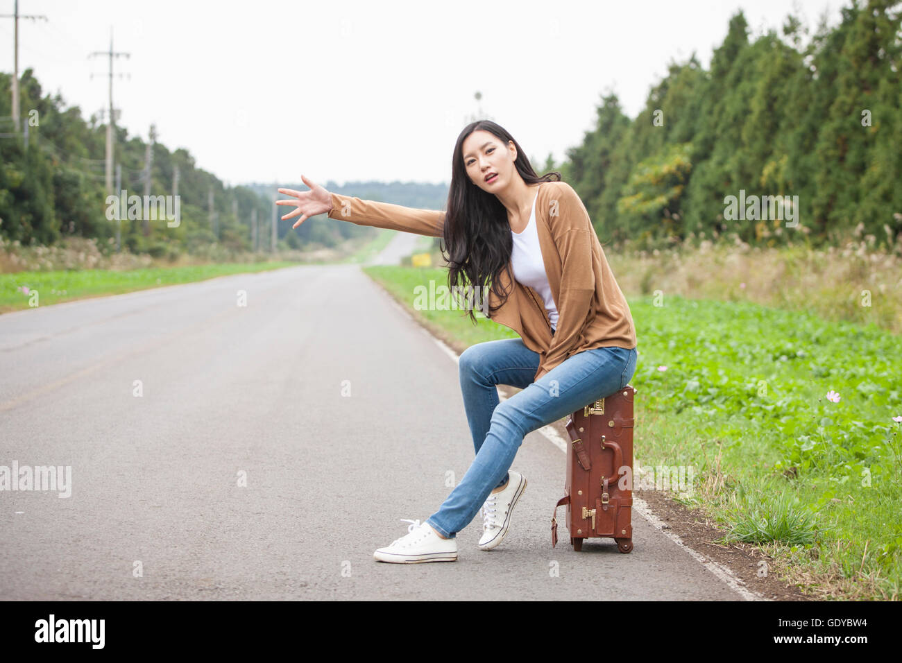 Junge Frau sitzt auf einem Träger und versucht, eine Mitfahrgelegenheit auf einer Straße Stockfoto