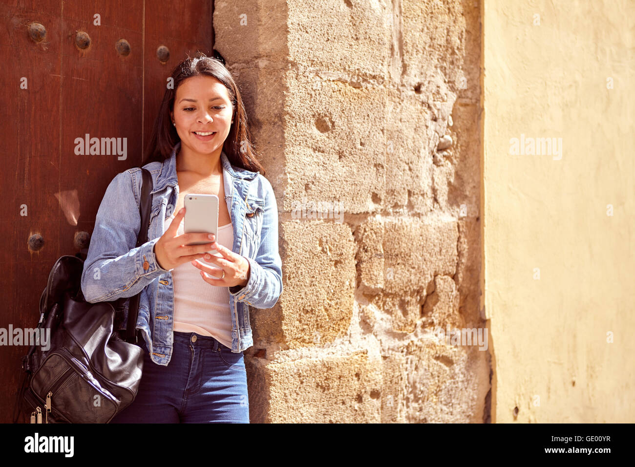 Hübsches junges Mädchen lehnte sich gegen einen alten Stahltür Blick auf ihr Handy beim Lächeln und ihr langes Haar locker tragen casua Stockfoto