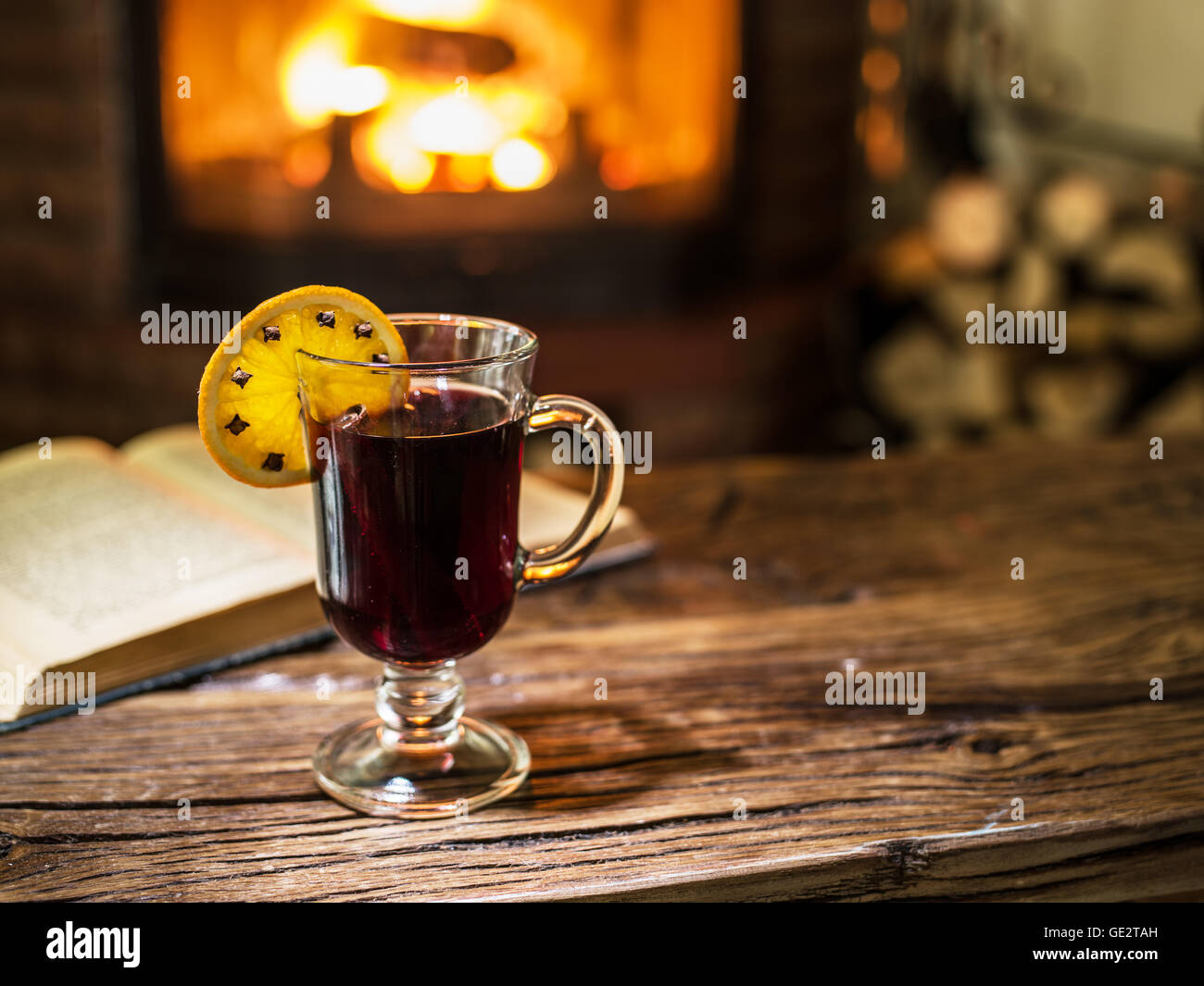 Glühwein und ein Buch auf dem Holztisch. Kamin mit warmen Feuer im Hintergrund. Stockfoto