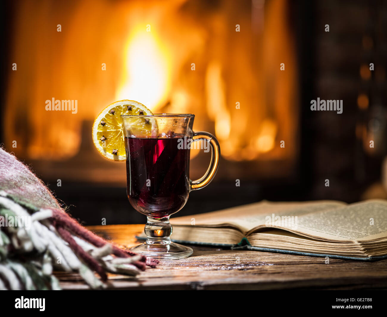 Glühwein und ein Buch auf dem Holztisch. Kamin mit warmen Feuer im Hintergrund. Stockfoto