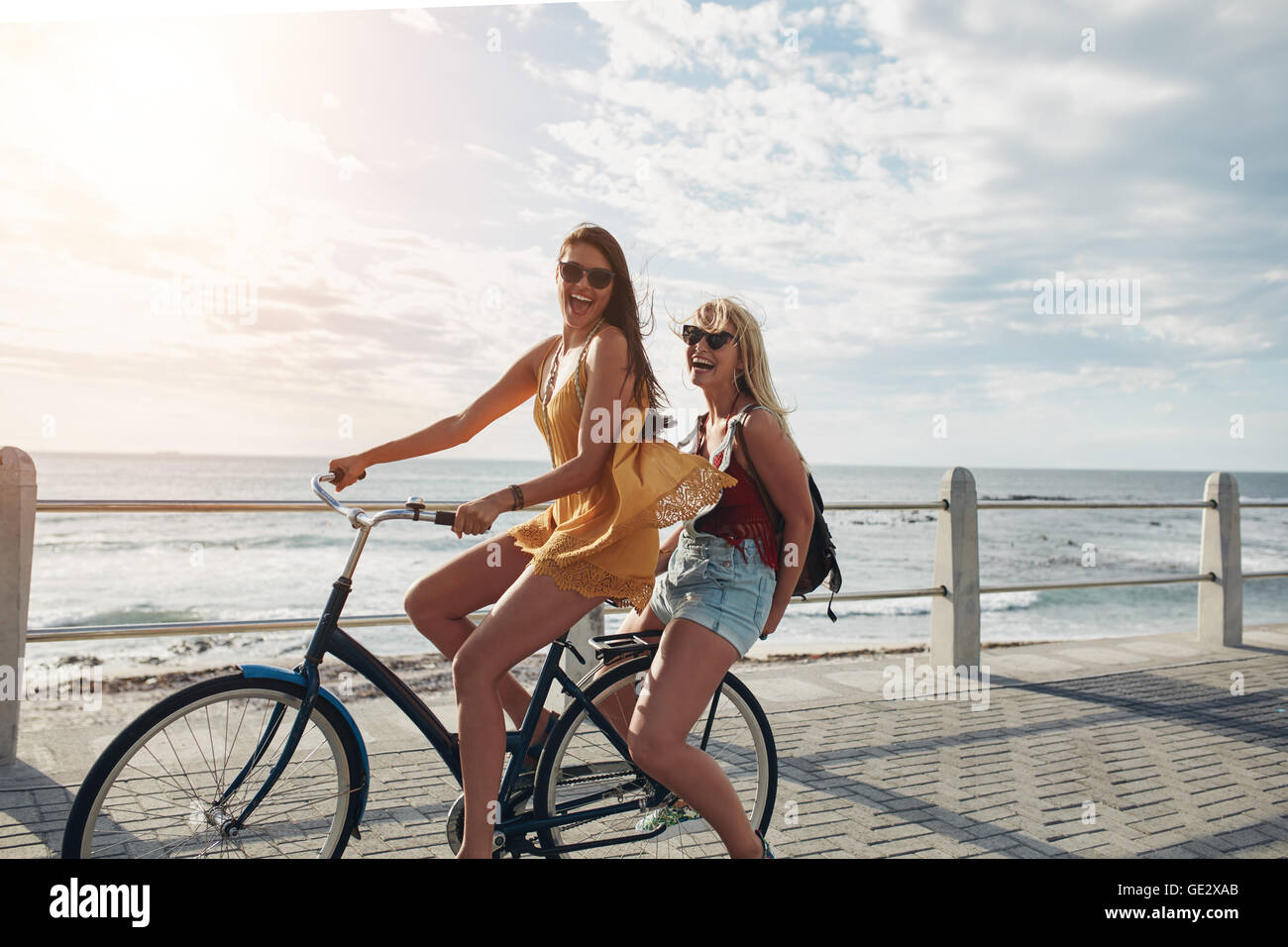 Zwei fröhliche junge Freundinnen auf einem Fahrrad. Beste Freunde genießen ein Fahrrad fahren an einem sonnigen Tag an der Uferpromenade. Stockfoto