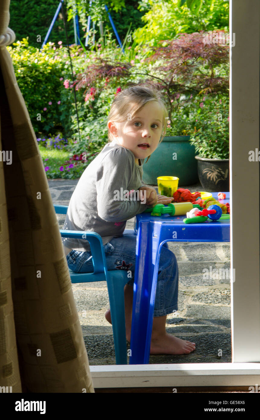 Drei Jahre alten Mädchen, das durch offene Terrassentür spielen mit Play-Doh Modellierung Kitt gesehen. VEREINIGTES KÖNIGREICH. Draußen im Garten. Stockfoto