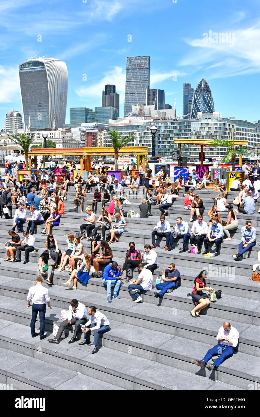 Büromitarbeiter machen Mittagspause bei heißem Sonnenschein im Amphitheater More London Scoop am Flussufer mit der Skyline der City of London jenseits von Southwark England UK Stockfoto