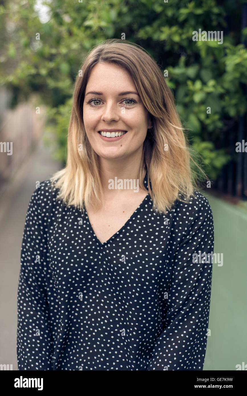 Profilbildnis einer jungen Frau mit viel Grün hinter ihr während lächelnd auf der Straße Stockfoto