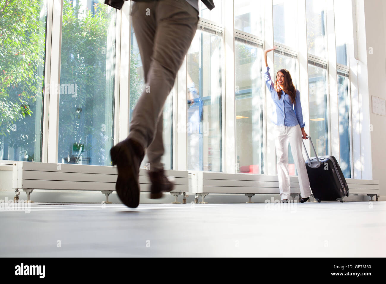 Glückliche junge Frau mit Gepäck zu ihrem Freund winken. Mann läuft in Richtung Partnerin am Flughafen. Sie ist mit Gepäck an de Stockfoto