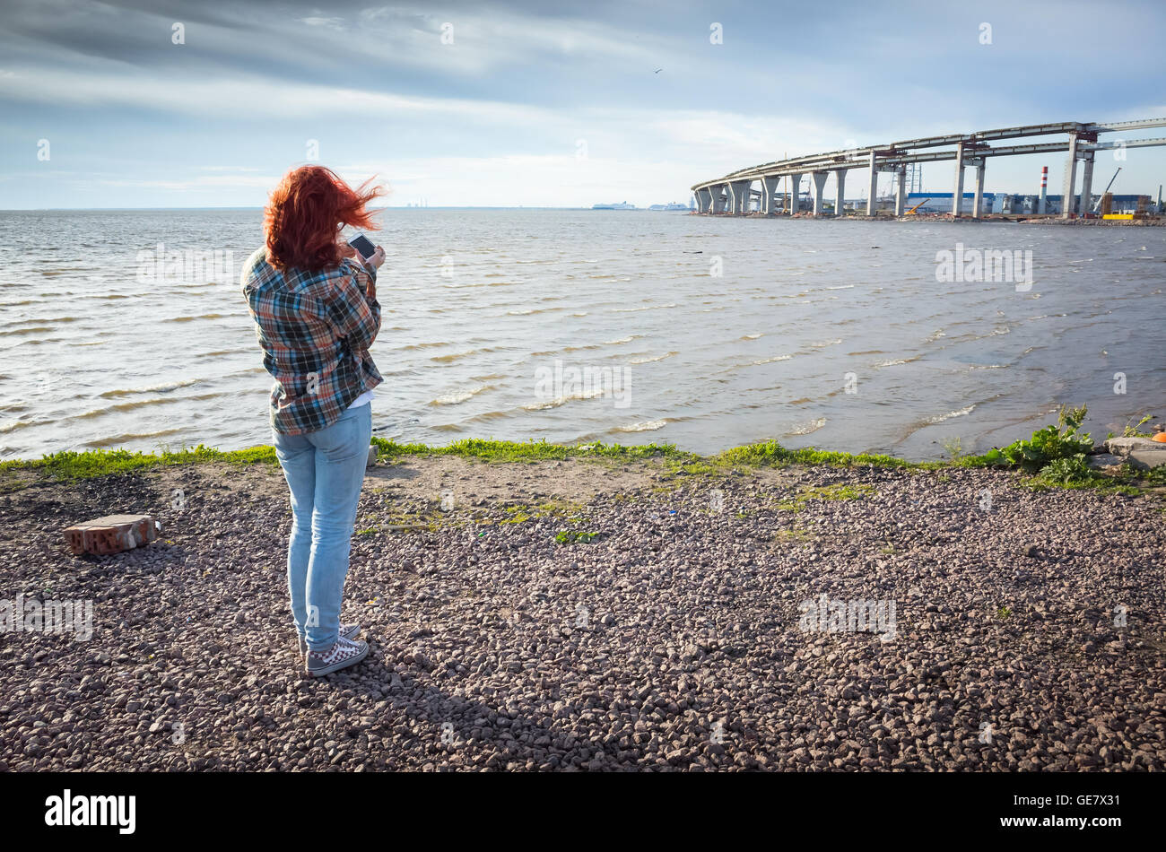 Teenager-Mädchen mit roten Haaren unter Telefon Landschaftsfoto mit modernen Brücke im Bau auf einer Küste Stockfoto