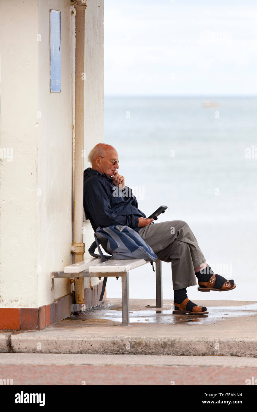 Llandudno, Wales, UK. 25. Juli 2016. UK Wetter – einem bewölkten Tag mit unter durchschnittliche Temperaturen an das Seebad Llandudno, Nordwales. Trotz des Wetters erfreuen Menschen immerhin das Resort. Ein Mann ein Buch an einem der vielen Strandpromenade Schutzhütten entlang der Promenade genießen Stockfoto