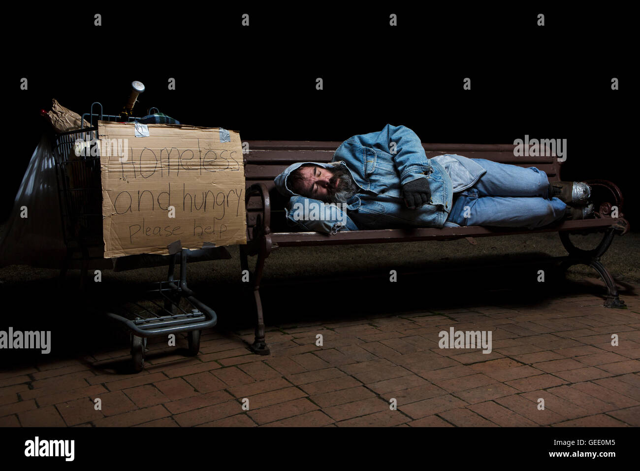 Obdachloser in der Nacht schlafen auf einer Parkbank mit seinem Einkaufswagen voller seinen Besitz Stockfoto