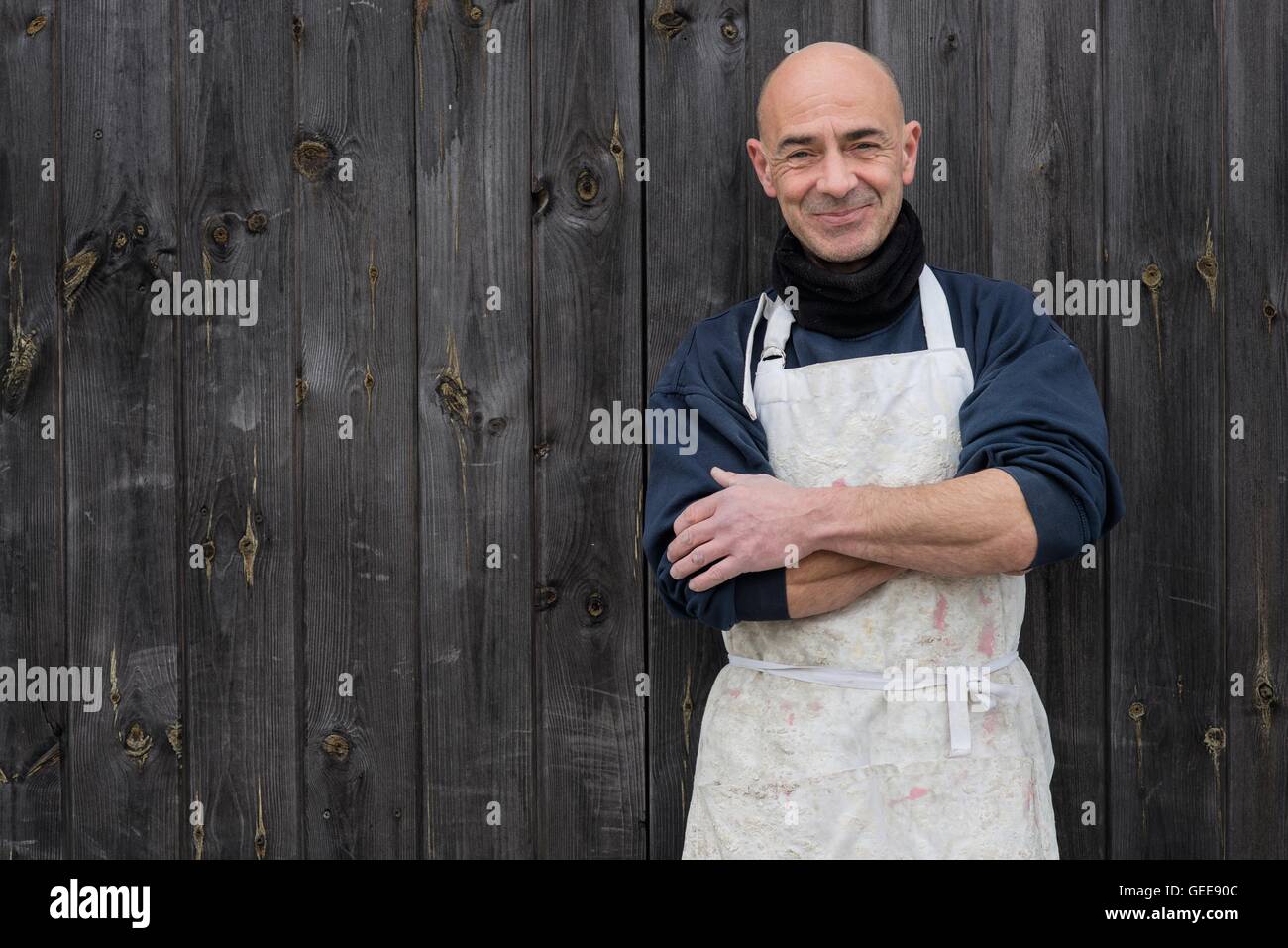 Kahlköpfiger Mann in dicke Kleidung und weiße Schürze lächelnd und mit verschränkten in eine Holzplatte Hintergrund stehend Stockfoto