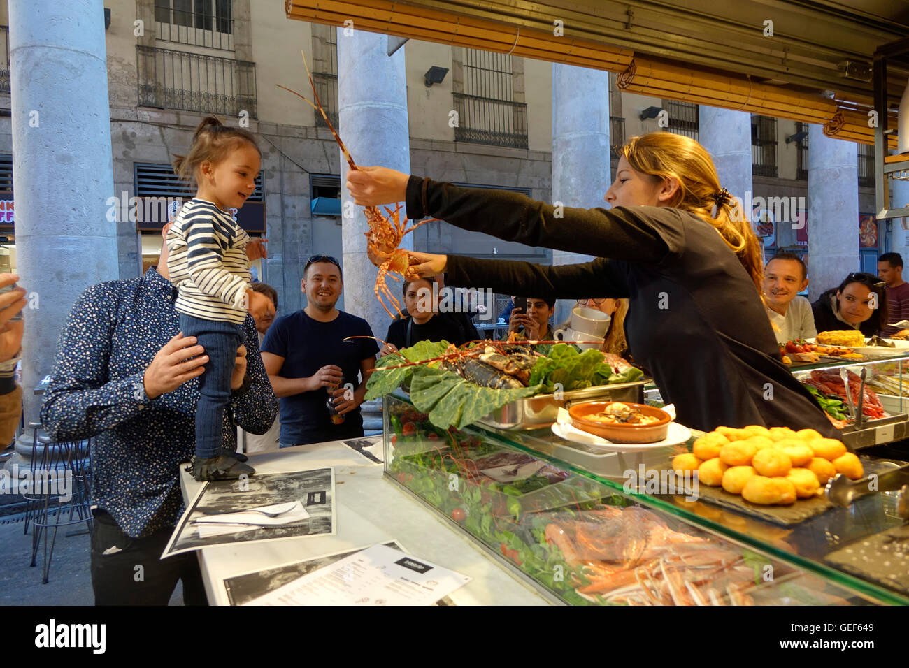 Ein kleines Mädchen wird von einer Kellnerin mit einem Hummer an einem Restaurant-Schalter auf dem La Boqueria-Markt in Barcelona unterhalten. Stockfoto