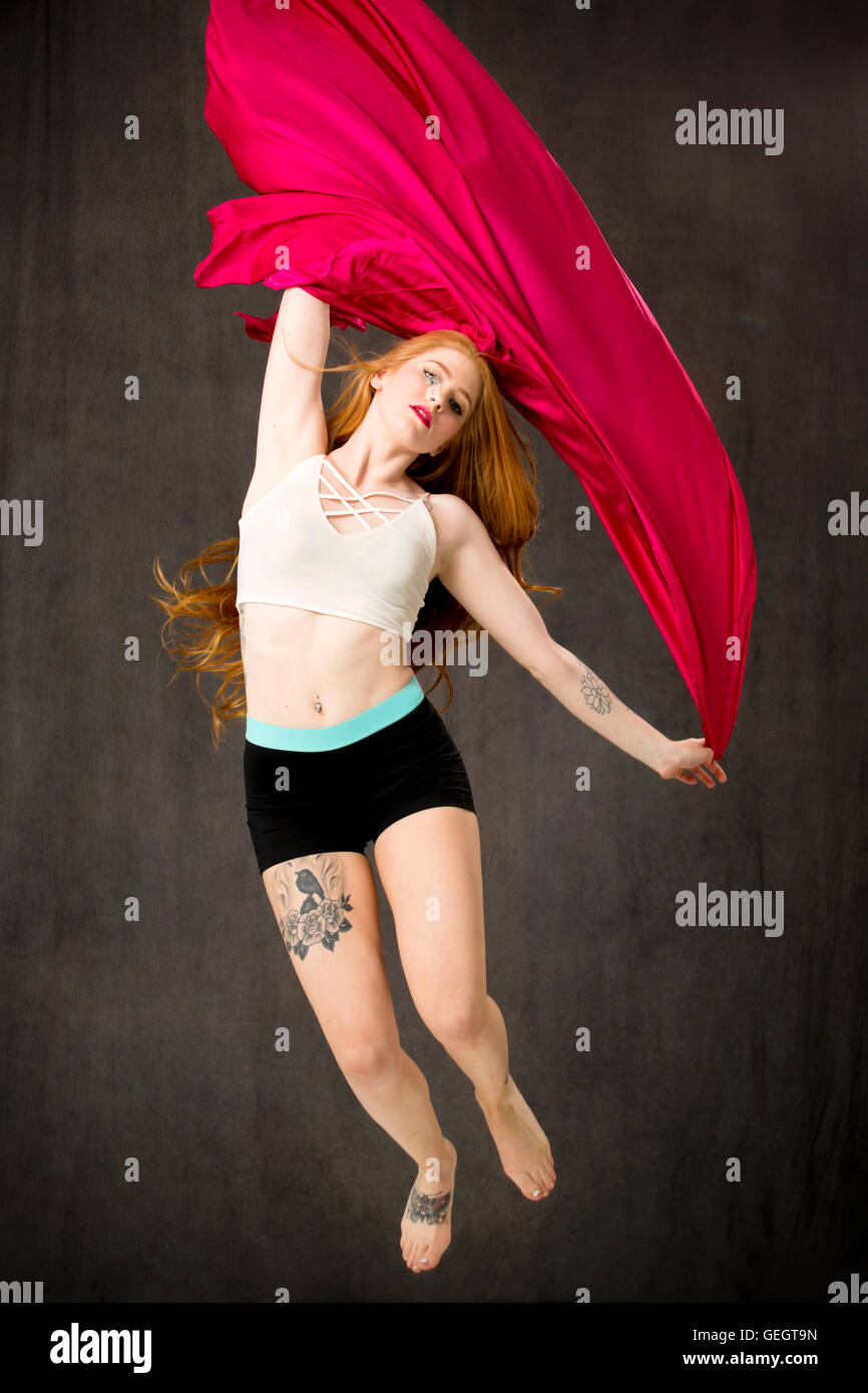 Junge Tänzerin mit langen roten Haaren und Tätowierungen mit rotem Stoff, der Bäche, während sie springen ist. Stockfoto
