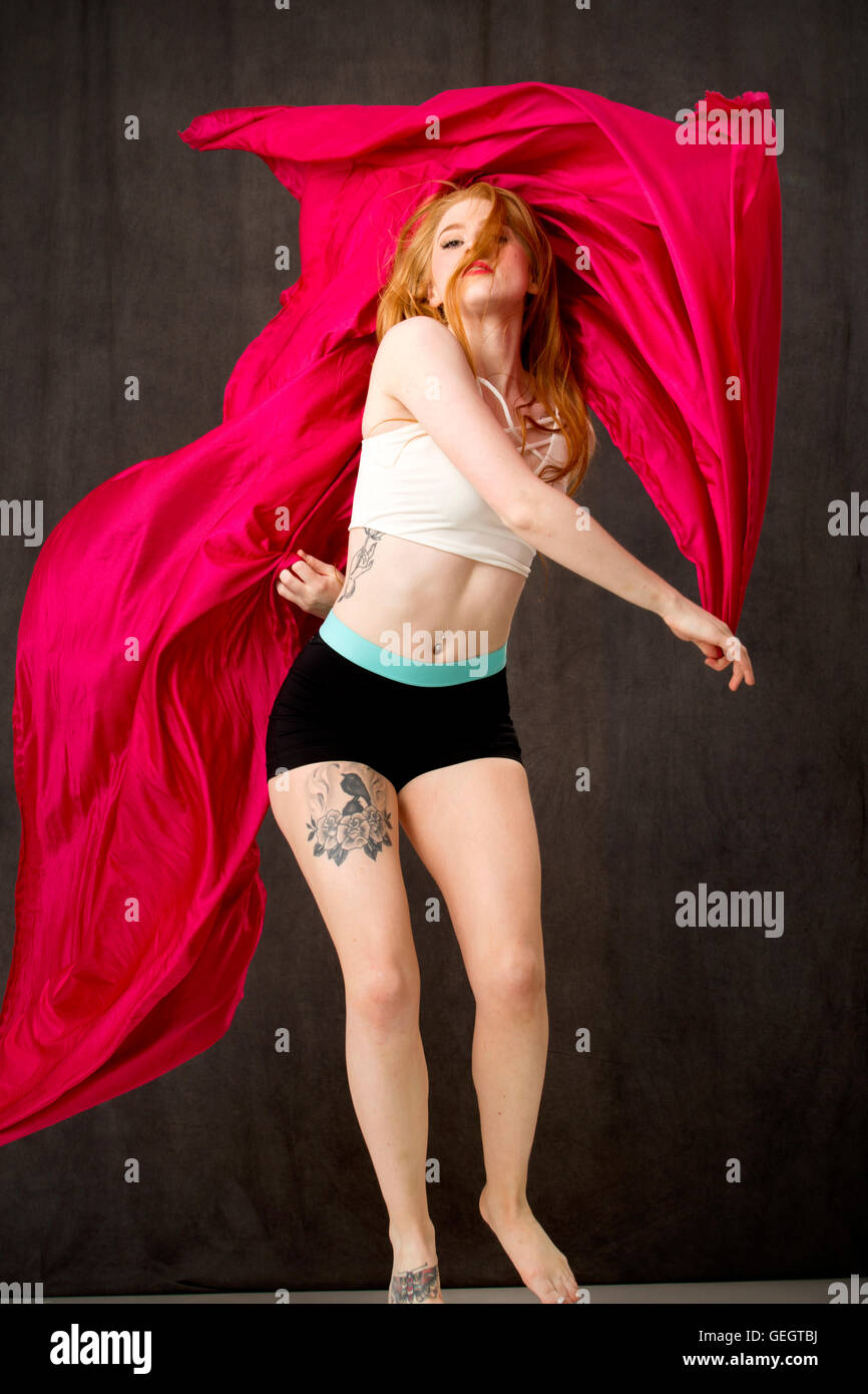 Junge Tänzerin mit langen roten Haaren und Tätowierungen mit rotem Stoff, der Bäche, während sie springen ist. Stockfoto