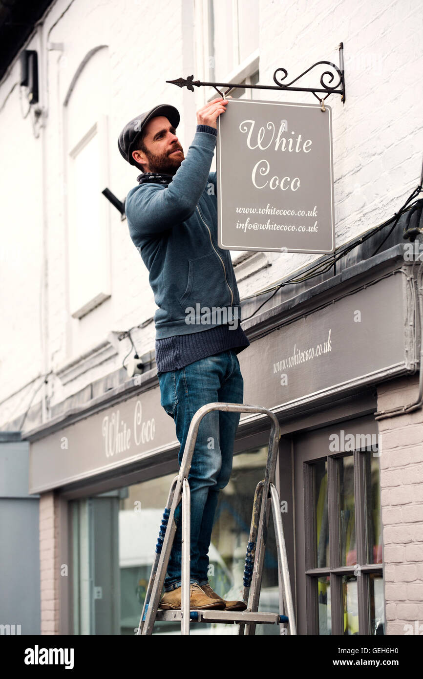 Ein Mann auf einer Leiter Befestigung eine gemalte Namensschild auf einer Halterung an einer Shops. Stockfoto